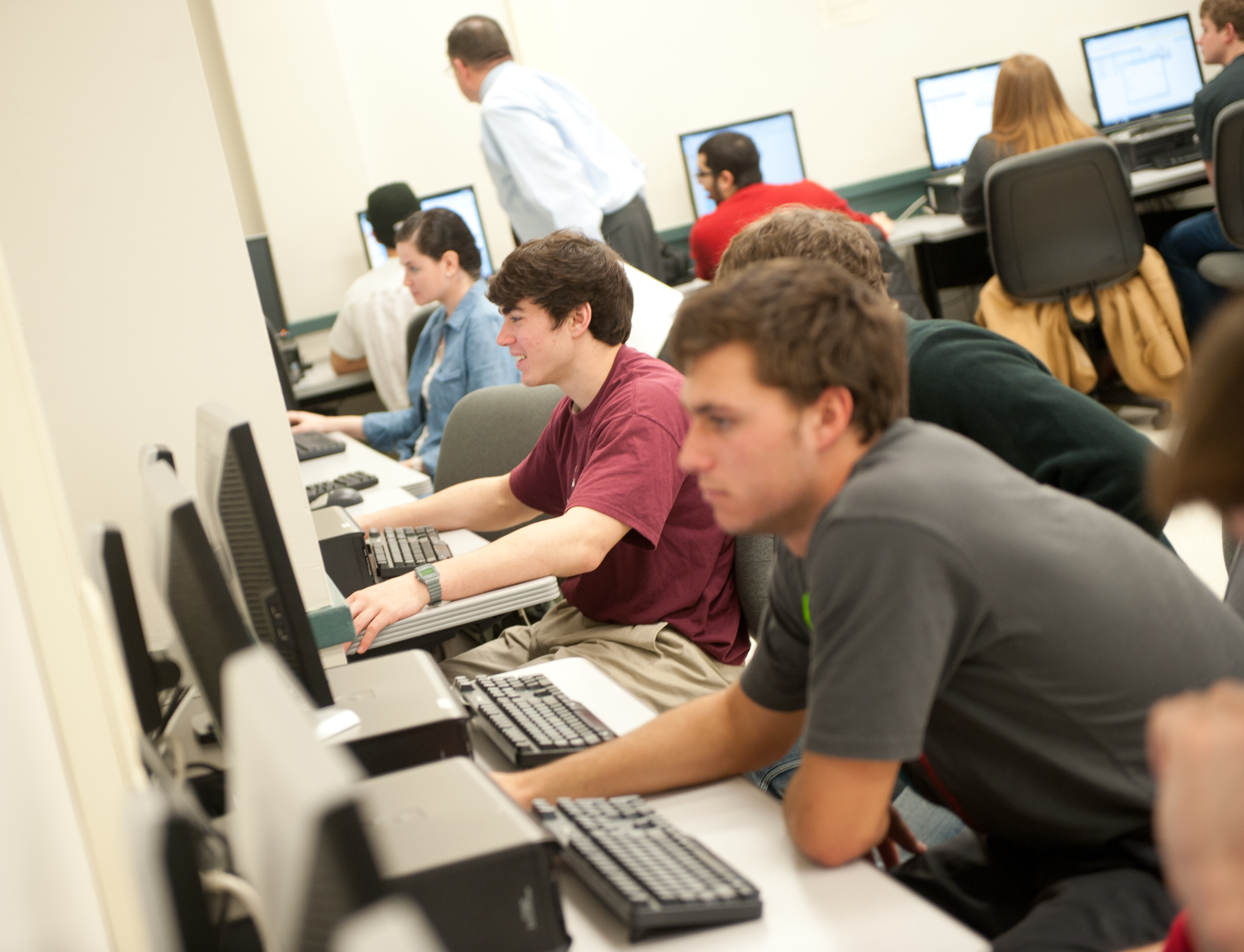 Students in a computer lab, with an instructor offering guidance to a student
